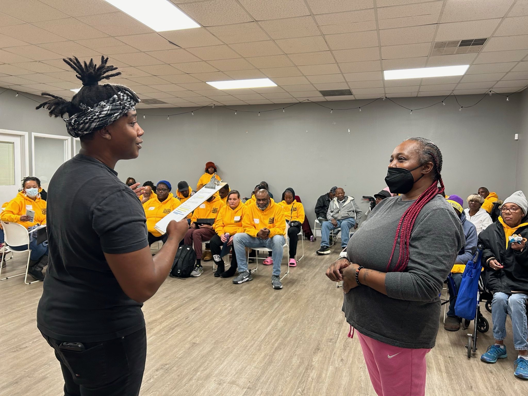 2 Black people speaking to each other in front of a crowd of Black & Brown people who have yellow hoodies on.